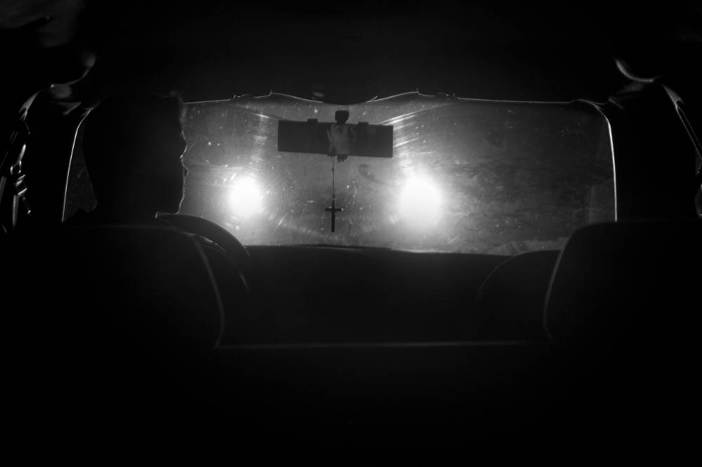 oncoming headlights silhouette crucifix hanging from rear view mirror in this black and white photo